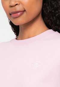 Sweatshirt roz deschis cu decolteu rotund și manșete rib. Prezintă inițialele brodate în alb pe partea stângă. Textură de țesătură moale.