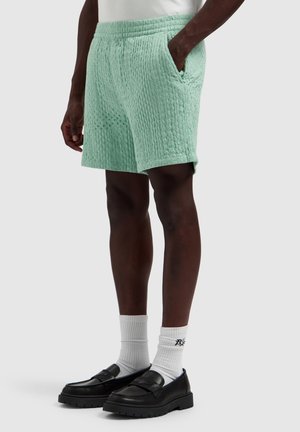Mann trägt strukturierte hellgrüne Shorts, weiße Socken und schwarze Loafers, steht mit einer Hand in der Tasche auf grauem Hintergrund.