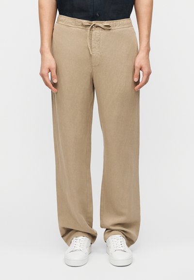 Personne portant un pantalon beige ample à cordon de serrage et des baskets blanches, debout devant un fond clair uni.