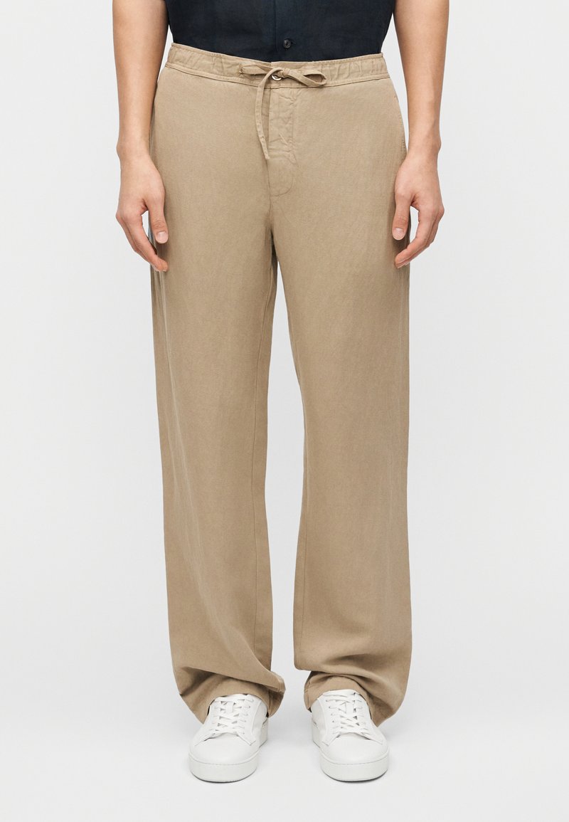 Άτομο που φορά χαλαρό μπεζ παντελό νι με κορδόνι και λευκά αθλητικά παπούτσια, στέκεται μπροστά σε μια απλή ανοιχτόχρωμη επιφάνεια.