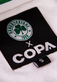 Witte stoffen label met een groen klavertje vier-logo met "Παναθηναϊκός 1908" en een zwart gedeelte waarop "x COPA" in witte letters staat. Maat S label zichtbaar.
