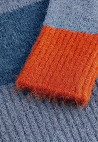 Brede wollen trui detail met een ribbelige oranjemouw tegen een gestructureerd blauwe body, voorzien van een fluweelachtige afwerking en zachte, gelaagde patronen.