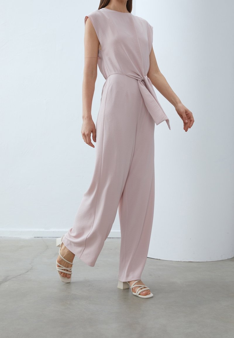 Femme marchant dans une combinaison sans manches rose clair avec une taille nouée, portant des sandales blanches à talons sur un sol gris.