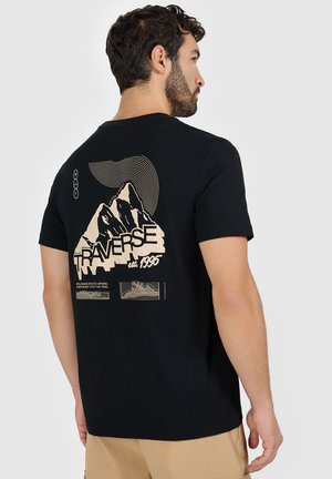 Mężczyzna w czarnej koszulce z beżową grafiką gór, "TRAVERSE est. 1995," oraz abstrakcyjnymi liniowymi wzorami na plecach, zwrócony tyłem.