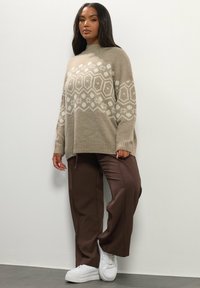 Beige gebreide trui met witte geometrische patronen, gecombineerd met bruine wijde broek en witte sneakers. Zachte textuur, ontspannen pasvorm.