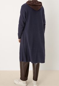 Langer, navyblauer, gerippter Cardigan mit einer braunen Kapuze, kombiniert mit braunen, faux Lederhosen und cremefarbenen Turnschuhen. Texturierte Stoffdetails sind sichtbar.