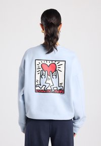 Lichtblauwe sweatshirt met een grote afbeelding op de achterkant van twee handen die een rood hart vasthouden, omlijnd in zwart met zonnestraaldetails.