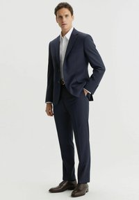 Granatowy garnitur o dopasowanym kroju, jednorzędowa marynarka, klapy w stylu notch oraz kieszenie boczne, połączony z białą koszulą i brązowymi butami.