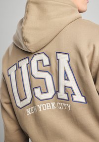 Tan hoodie featuring large embroidered "USA" in white outlined with blue and "NEW YORK CITY" in smaller white text below. Soft texture.