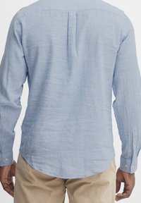 Maglietta a maniche lunghe azzurro chiaro realizzata in un tessuto texturizzato, con una piega sul retro e orlo arrotondato. Polsini con bottoni di colore verde chiaro.