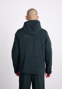 Mörkgrön fleecehuva med stor huva, långa ärmar och avslappnad passform. Slät textur och minimalistisk design.