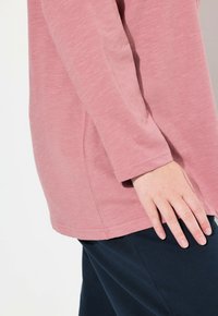 Top rosa chiaro a maniche lunghe con una texture liscia, spacco laterale e dettagli di cucitura sottili, abbinato a pantaloni scuri.