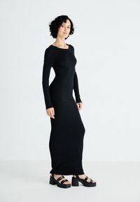 Vestido negro de manga larga con textura, de silueta ajustada y cuello redondo, combinado con sandalias de plataforma negras y gruesas.