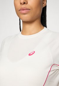 Nærbillede af en person iført en hvid tekstureret sports skjorte med pink detaljer og et pink ASICS-logo på brystet.