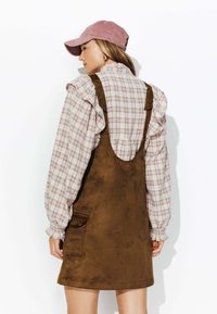 Robe en suede marron avec bretelles, associée à une chemise à manches longues à carreaux clairs et une casquette en velours côtelé rose ; la vue de dos montre des manches volantes.