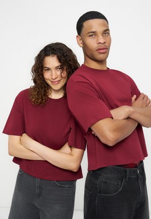 To unge voksne iført matchende vinrøde T-shirts og sorte jeans står ryg mod ryg med armene krydset og smiler blødt og selvsikkert.