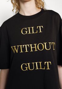 Černé bavlněné tričko s odvážným zlatým vyšitým textem, který zní "GILT WITHOUT GUILT" ve třech řádcích. Kulatý výstřih, krátké rukávy.