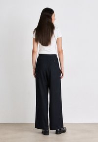 Pantaloni a gamba larga in tessuto a righe sottili scure, con vita alta, tasche posteriori e una finitura liscia e testurizzata. Indossati con una semplice t-shirt bianca.