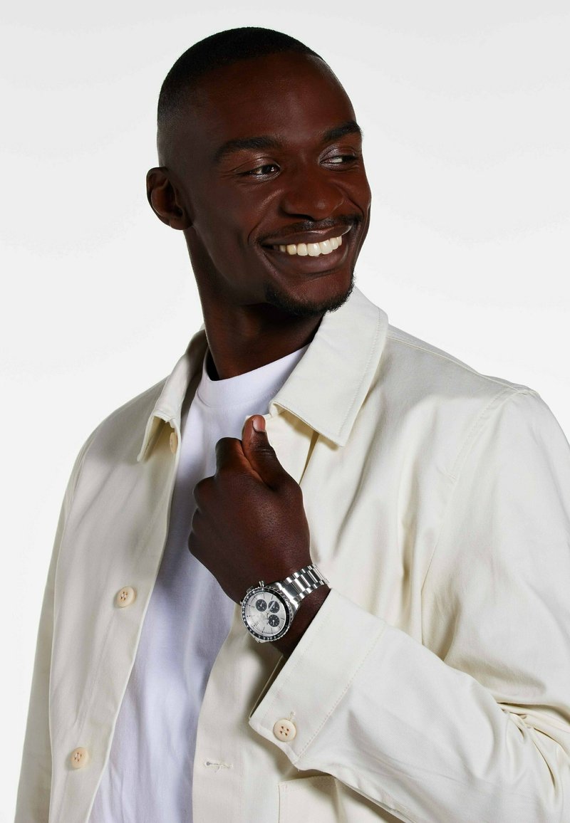 Silver watch with a textured white face and black dials. Model wears a cream jacket and white shirt. Skin tone is deep brown.