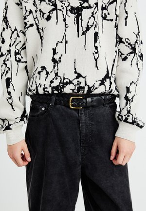 Cinturón de cuero trenzado negro con hebilla dorada, llevado sobre pantalones de pana negros y holgados. El suéter presenta una tela blanca con un patrón floral abstracto en negro.