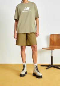 Olivově zelené přessize tričko s bílým logem, zkombinované s khaki šortkami. Model má na sobě světle šedé šněrovací boty s černou podrážkou.