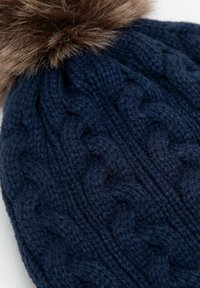 Tmavě modrá pletená čepice s copánkovým vzorem a hnědým umělým kožešinovým bambulí na vrchu. Materiál působí silně a teple, s texturovaným povrchem.