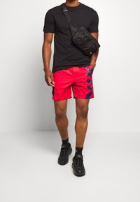 Shorts rouges avec des accents navy et un motif en chevrons, associés à un t-shirt noir et des baskets noires. La tenue comprend un sac à bandoulière noir.