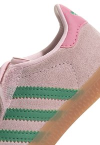 Light pink suede sneaker with green zigzag stripes and a pink heel accent. Features a semi-transparent gum sole for traction.