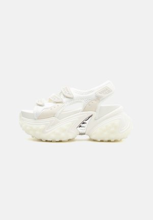 Sandale de sport blanche épaisse avec des brides Velcro ajustables, des détails perforés, une semelle épaisse texturée et un accent argenté sur la voûte plantaire.