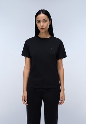 Femme aux longs cheveux noirs portant un t-shirt noir uni à manches courtes et un pantalon noir, debout devant un fond gris clair.