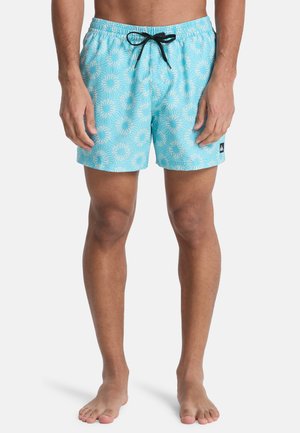Mann trägt hellblaue Schwimmshorts mit weißen Sonnenstrahlenmustern und einem schwarzen Kordelzug vor weißem Hintergrund.
