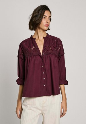 Burgunderfarbene Bluse mit einer Rüschenspitze und floralen Durchbrüchen, mit Knopfverschluss und aufgerollten Ärmeln. Aus weichem Baumwollmaterial.