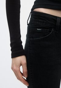 Pantalones vaqueros negros de denim con un ajuste entallado, que cuentan con un diseño clásico de cinco bolsillos y una pequeña etiqueta con la marca en el bolsillo delantero. La textura es suave.