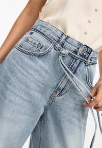 Persona vistiendo jeans de mezclilla de talle alto en azul claro con un cinturón atado y una blusa cremoso con botones, mano sosteniendo el extremo del cinturón.
