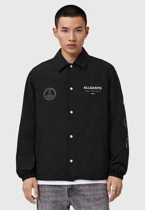 Black jacket with snap buttons, peace symbol embroidery on chest and sleeve, elastic cuffs, and a smooth texture. Brand name visible.