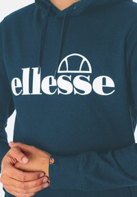 Navyblaue Kapuzenjacke aus Baumwolle mit einem großen weißen "ellesse"-Logo und einem Emblem an der Manschette. Sie verfügt über eine Kordelzugkapuze und gerippte Manschetten.