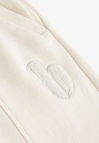 Cream-colored sweatshirt fabric with a soft texture. Features a raised, textured '10' embroidered detail near the shoulder seam.