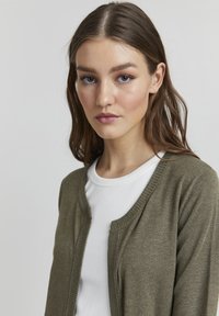 Olive green cardigan with a rounded neckline and thin, ribbed edges, worn over a white shirt. Soft texture and simple design.