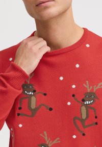 Czerwony sweter dzianinowy z wzorem renifera, z białymi akcentami w postaci płatków śniegu oraz okrągłym dekoltem. Textura jest gładka z ściągaczami na mankietach.