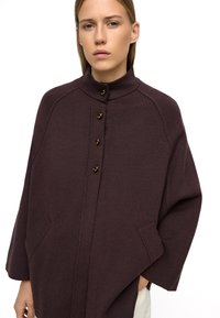 Brązowy sweter z wysokim kołnierzem, z złotymi guzikami i bocznymi kieszeniami, zaprojektowany w luźnym kroju z minimalną ilością szwów.