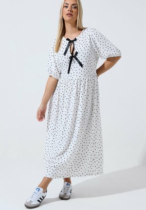 Kvinde iført hvid midi-kjole med sorte polkaprikker, pufærmer, to sorte båndsløjfer foran og hvide sneakers med sorte striber.