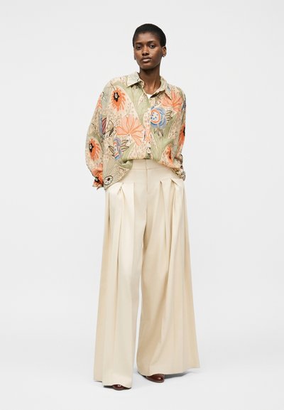 Femme portant un chemisier à imprimé floral ample avec des fleurs orange et bleues, associé à un pantalon crème à taille haute et jambes larges, ainsi qu'à des chaussures marron.