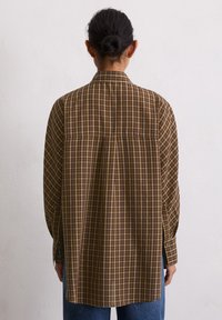 Chemise décontractée à carreaux marron avec des manches longues, des poignets boutonnés et un ourlet arrondi, contrastée avec un jean en denim clair.