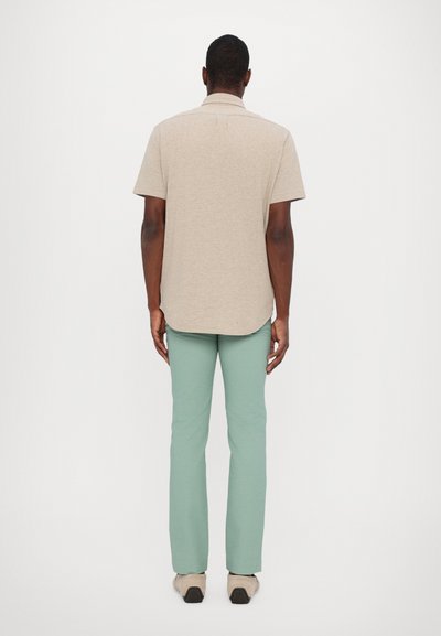 Camisa de manga corta color beige con cuello, combinada con pantalones verdes claros, vista desde atrás. Material de algodón, textura suave, diseño sencillo.