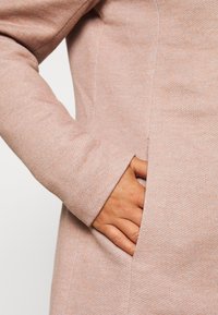 Cappotto rosa chiaro caratterizzato da un tessuto texture, con una tasca laterale e maniche aderenti. Il materiale sembra morbido e comodo.