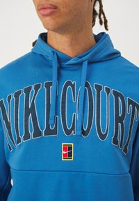 Sudo de capuz azul feito de tecido macio apresenta grande lettering "NIKELCOURT" e um pequeno emblema da Nike, com cordão no capuz.