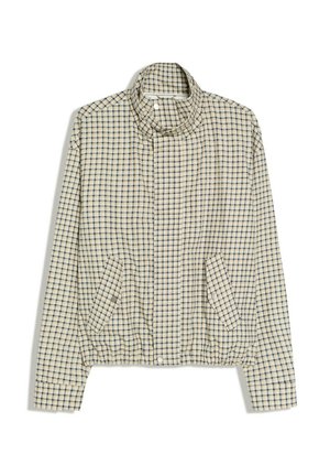 Mand iført mørke solbriller, beige ternet jakke, blå skjorte, sort slips og brede sorte bukser står indendørs ved en betonpille.