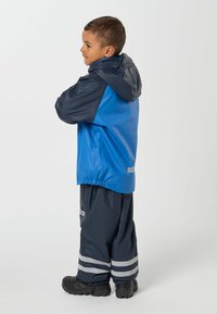 Jeune garçon debout, les bras croisés, portant une veste et un pantalon imperméables bleu et marine avec des bandes réfléchissantes, bottes noires.