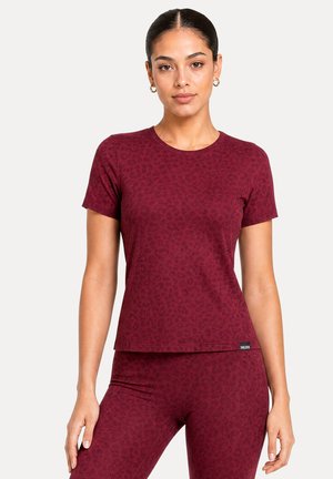 Femme portant un haut ajusté bordeaux à manches courtes et des leggings assortis avec un motif léopard subtil, debout devant un fond uni.