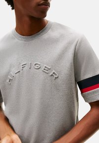 Camiseta de algodón gris con cuello redondo, texto bordado "H.L. FINGER" y acentos a rayas en azul y rojo en las mangas.